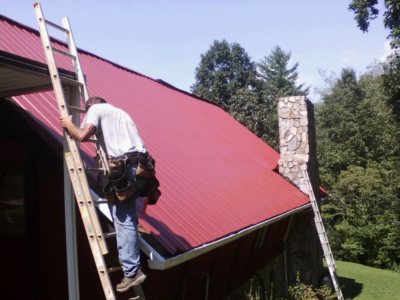 Roofer on a ladder beside a red metal roof and stone chimney — Soffit & Fascia Repair by Roofing Brothers in Rufe